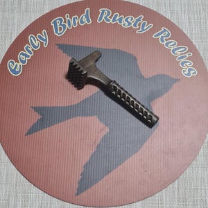 May include: A dark, textured meat tenderizer hammer with a patterned handle rests on a round, red and blue surface. The surface features a bird silhouette and the text "Early Bird Rusty Relics" in a blue, cursive font.