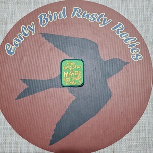 May include: A circular design with the text "Early Bird Rusty Relics" in light blue. A dark gray bird silhouette is centered on a reddish-brown background. A green patch with the text "the decision is MAYBE and that's FINAL!!" is in the center.