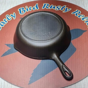 May include: A black cast iron skillet with a handle, labeled "NO. 5 SKIN." The skillet is centered on a round, red and brown background with the text "Early Bird Rusty Relics" in blue. The skillet is a kitchenware item.