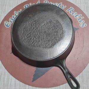 May include: A black cast iron skillet with a long handle. The skillet has a textured cooking surface and the words "WAGNER" and "SIDNEY O." are embossed on the bottom. The skillet is on a red and brown surface with the words "Early Bird Dusty Relics".
