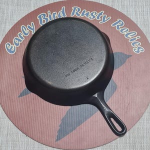 May include: A black cast iron skillet with a long handle, resting on a round, pink and blue patterned surface. The skillet has the text "995 IRON SKILLET" on its base. The surface has the text "Early Bird Rusty Relics".