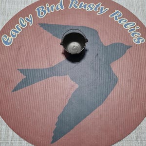 May include: A small, dark metal bucket with a handle sits on a round, reddish-brown surface. The surface features a silhouette of a bird in gray and the text "Early Bird Rusty Relics" in blue. The bucket is likely vintage.