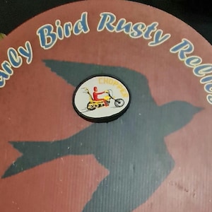 May include: A circular design with the text "Early Bird Rusty Relics" in blue and white. A black bird silhouette is visible on a reddish-brown background. A small patch shows a person riding a yellow motorcycle.