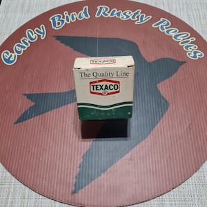May include: A small, vintage Texaco box with the words "The Quality Line" printed on the front. The box is white with green and red accents and the Texaco logo. The box is on a round, red and brown background with the words "Early Bird Rusty Relics".