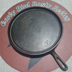 May include: A black cast iron griddle pan with a handle. The pan has a circular cooking surface and is marked with the text "Wagner Ware Sidney -O-" and the numbers "1109 E". The pan is set on a round sign with the text "Early Bird Rusty Relics".