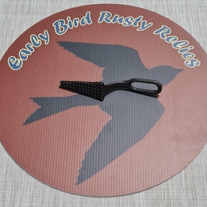 May include: A black metal tool with a textured, triangular blade and a looped handle rests on a round, red and gray patterned surface. The surface features a bird silhouette and the text "Early Bird Rusty Relics" in blue.