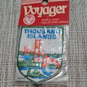 May include: A white embroidered patch with a red bridge and green trees. The patch is shaped like a shield and has the text "Thousand Islands" in blue lettering. The patch is packaged in a clear plastic bag with a red and white label that says "Voyager World Wide Collectors Series".