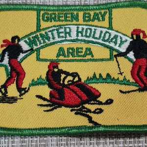 May include: A yellow embroidered patch with green trim featuring the text "Green Bay Winter Holiday Area" with two figures on skis and a snowmobile.