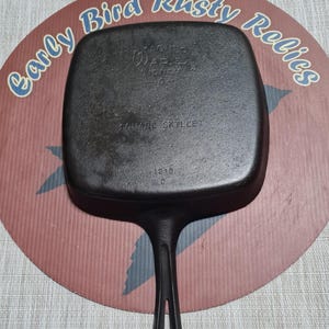 May include: A black square Wagner Ware Sidney O cast iron skillet with a long handle. The skillet is marked "SQUARE SKILLET" and "1218 0". The skillet is on a round sign that says "Early Bird Rusty Relics".