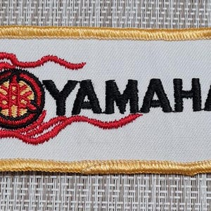 May include: A white embroidered patch with a gold border. The patch features a red and black stylized logo with a flame design and the word "YAMAHA" in black.