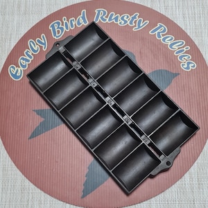 May include: A black cast iron muffin pan with 12 individual molds. The pan is rectangular with rounded compartments and a central divider. The pan is on a red and pink background with the text "Early Bird Rusty Relics".
