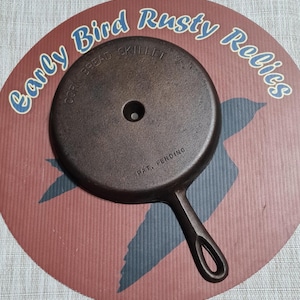 May include: A small, dark brown cast iron skillet with a handle and a circular design. The skillet is marked "Corn Bread Skillet" and "Pat. Pending." The skillet is on a round, red and blue background with the text "Early Bird Rusty Relics."