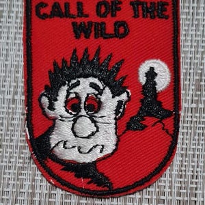 May include: Red embroidered patch with a black outline. The patch features a cartoon character with a white face and black hair, looking up at a white moon. The text "CALL OF THE WILD" is embroidered in black on the patch.