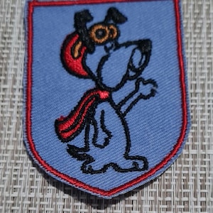May include: A blue embroidered patch with a red border, featuring a cartoon dog wearing a red cape and goggles. The dog is standing on its hind legs with its front paws raised.