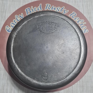 May include: A cast iron skillet lid with a dark gray finish. The lid is round with a raised edge and the number 9 is embossed on the bottom. The words "FAVORITE PIQUA WARE" are also embossed on the lid.