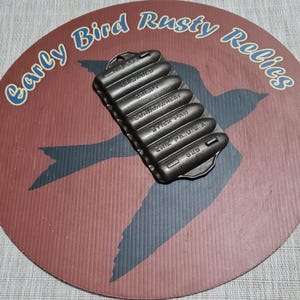 May include: A cast iron stick pan with the text "ERIE PA, U.S.A." and "625" on a red and black patterned background. The pan has several compartments and is labeled "CRISPY" and "CORN/WHEAT". The background has the text "Early Bird Rusty Relics".