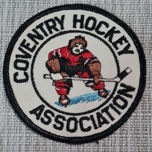 May include: A white embroidered patch with black stitching. The patch features a hockey player in a red and black jersey with a white maple leaf on the front. The text "Coventry Hockey Association" is written in a black circular border around the player.
