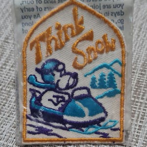 May include: Embroidered patch with the words "Think Snow" in orange script. The patch features a cartoon character on a snowmobile, with blue and purple accents, and a mountain scene. The patch is in a clear plastic sleeve.