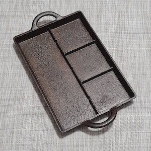 May include: A cast iron baking pan with four sections, each with a slightly raised edge. The pan has two handles on the sides and is dark brown in color.