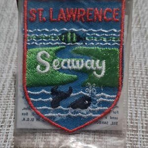 May include: A blue and green embroidered patch with the text "St. Lawrence Seaway" in white. The patch features a stylized image of a river with a fish and a seal.