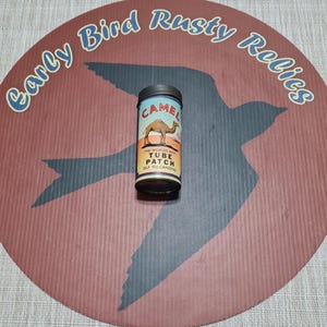 May include: A vintage Camel tube patch tin with a camel graphic and text. The tin is cylindrical with a black lid and base. The background is a circular design with a bird silhouette and text that reads "Early Bird Rusty Relics".