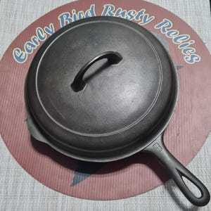 May include: A black cast iron skillet with a lid and handle. The skillet is sitting on a red and white circular surface with the text "Early Bird Rusty Relics".
