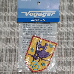 Peut inclure: Un écusson brodé jaune et violet représentant un couple dansant. L'écusson est en forme de bouclier avec une bordure rouge et des accents bleus. Le texte "Voyager originals" est imprimé sur l'emballage.
