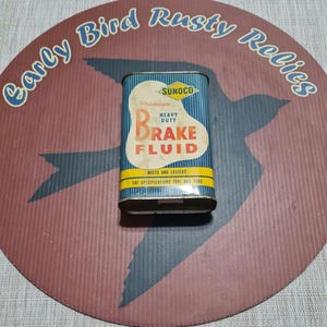 May include: Vintage Sunoco Premium Heavy Duty Brake Fluid tin. The rectangular tin has a blue and white striped background with the words "Brake Fluid" in a large, stylized font. The tin is on a circular background with the words "Early Bird Rusty Relics".