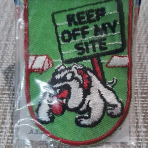 May include: A green embroidered patch with a white bulldog holding a red sign that says "Keep Off My Site".