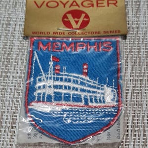 May include: A blue and red embroidered patch with the word "Memphis" in white lettering. The patch features a white steamboat on a blue body of water. The patch is packaged in a clear plastic bag with a Voyager World Wide Collectors Series label.