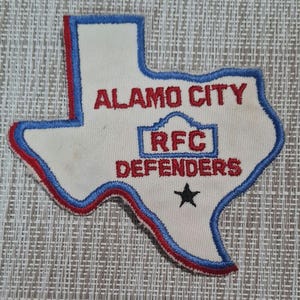 May include: A white patch shaped like the state of Texas with blue and red outlines. The patch has the text "Alamo City RFC Defenders" in red and a black star in the bottom center.