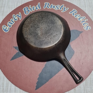 May include: A vintage cast iron skillet, approximately 8 inches in diameter, with a long handle and a circular cooking surface. The skillet is dark brown and sits on a red and blue patterned surface with the text "Early Bird Rusty Relics".