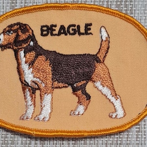 May include: An embroidered patch with a beige background and an orange border. The patch features a Beagle dog with brown and white fur. The word "BEAGLE" is embroidered in orange above the dog.