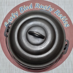 May include: A dark gray cast iron Dutch oven with a lid and a handle. The lid has a circular design and a metal handle. The pot has a metal handle on each side. The text "Early Bird Rusty Relics" is printed on the background.