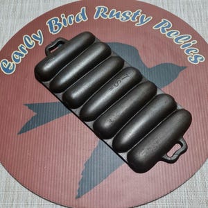 May include: A cast iron cornbread pan with six individual molds. The pan is dark gray and has handles on each end. The pan is sitting on a round, red and brown surface with the text "Early Bird Rusty Relics".