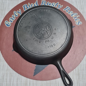 May include: A cast iron skillet with a handle, featuring the embossed text "GRISWOLD ERIE PA U.S.A." and "710 B". The skillet is dark gray and sits on a red and pink circular surface with the text "Early Bird Rusty Relics".