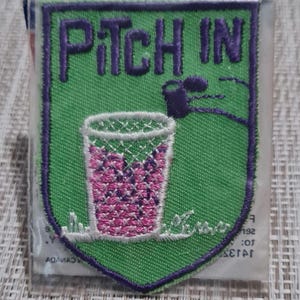 May include: Green felt patch with purple stitching and the words "Pitch In" in purple thread. A cartoon trash can with a pink and white pattern is in the center of the patch.