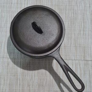 May include: A black cast iron skillet with a lid. The skillet has a long handle with a hole for hanging. The lid has a small handle. The skillet is sitting on a light gray surface.