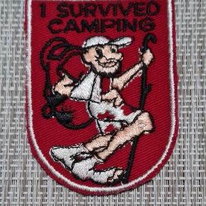 May include: Red embroidered patch with a white border. The patch features a cartoon character with a backpack and a walking stick. The text "I SURVIVED CAMPING" is above the character.