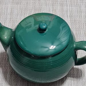 May include: A green ceramic teapot with a matching lid and handle. The teapot has a rounded body with horizontal ridges and a spout. The lid has a small knob on top. The teapot is approximately 7 inches wide and 6 inches tall.