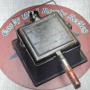 May include: A vintage cast iron square waffle iron with a wooden handle and a wire bail. The surface is embossed with the text: THE WAGNER MFG. CO. SIDNEY O. SQUARE WAFFLE IRON PAT'D FEB. 22 1910. The metal has a dark, weathered patina finish.