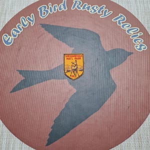 May include: A round, brown sign with the text "Early Bird Rusty Relics" in blue script. A dark blue bird silhouette is centered on the sign, with a small, yellow and orange emblem of Shenandoah Nat'l Park in the center.