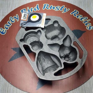 May include: A grey cast iron cake pan featuring four distinct character-shaped molds. The pan includes two integrated side handles and a rectangular paper tag attached to the top, displaying the Lodge logo and a graphic of a fried egg.