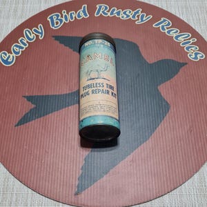 Puede incluir: Kit de reparación de tapones para neumáticos sin cámara Camel vintage en un recipiente metálico cilíndrico. El recipiente es azul claro con el logotipo y el texto de Camel. El fondo es un letrero circular con texto y una silueta de pájaro.