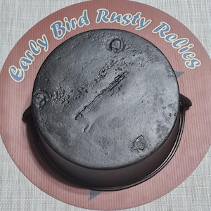 May include: A black cast iron pot with a handle, sitting on a round, pink and brown surface. The pot has a textured surface and a lid. The text "Early Bird Rusty Relics" is printed on the surface in blue.