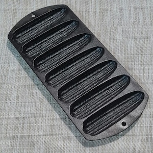 May include: Black cast iron cornbread pan with eight individual molds shaped like corn cobs. The pan has a smooth, slightly textured surface.