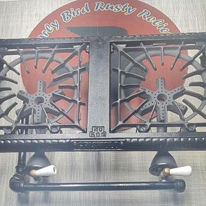 May include: A black cast iron double burner stove with a rectangular frame, two star-shaped burner heads, and four legs. The front features the text "GRISWOLD" and "NO 502" embossed on the metal. Two white ceramic handles are attached to the gas valves.