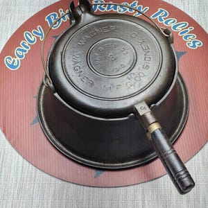 May include: A dark gray cast iron waffle maker with a wire handle and a wooden grip. The lid features embossed text reading WAGNER MFG CO SIDNEY O, PATD FEB 22 1910, and the number 8. The circular metal base has a dark, weathered, metallic finish.