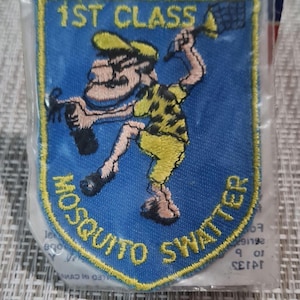May include: A blue and yellow embroidered patch with a cartoon character holding a fly swatter. The text "1st Class Mosquito Swatter" is embroidered around the character.