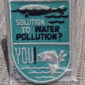 May include: A blue and white embroidered patch with the text "Solution to Water Pollution? You!" and a fish jumping out of the water. The patch is surrounded by a white border.